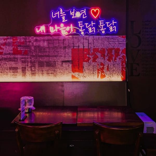 Neon Korean script and a heart sign glow above a dark wooden table and chairs at Mr. Dak.