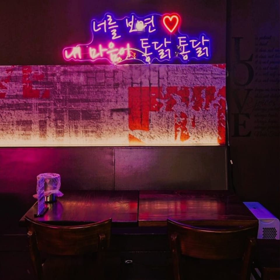 Neon Korean script and a heart sign glow above a dark wooden table and chairs at Mr. Dak.