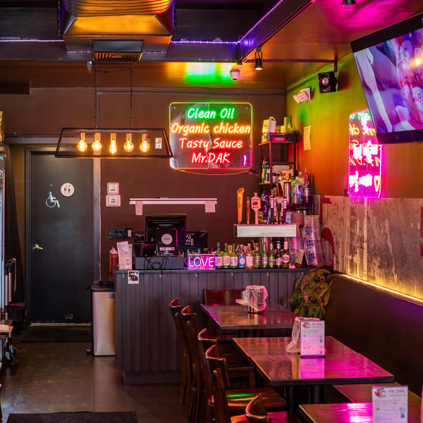 Neon-lit interior of Mr. Dak restaurant with tables, chairs, a bar, and a "Clean Oil" sign.