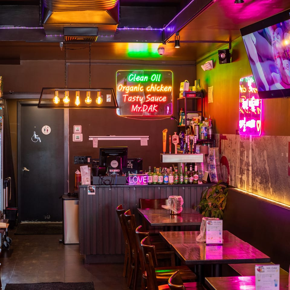 Neon-lit interior of Mr. Dak restaurant with tables, chairs, a bar, and a "Clean Oil" sign.