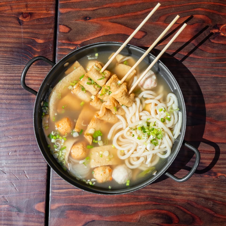 Overhead view of a hot pot with Korean fish cakes, udon noodles, and green onions.